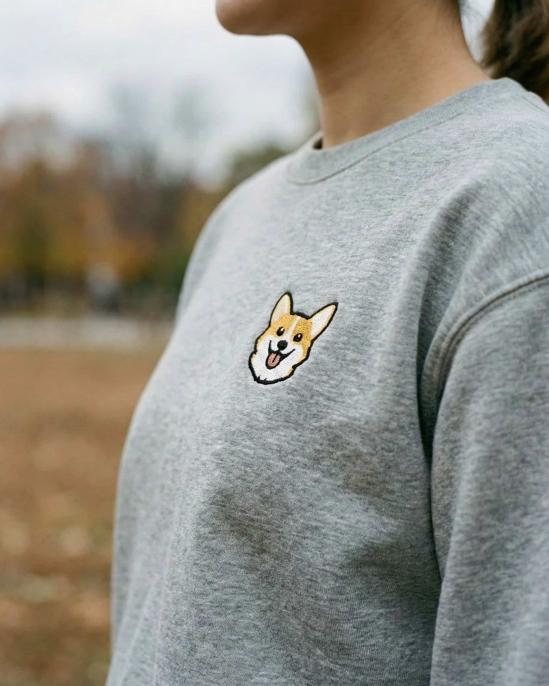 Woman wearing Light Grey Heather Sweatshirt with minimalist embroidered Corgi icon on left chest.