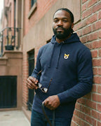 Man leaning against building wall, wearing Navy Hoodie with minimalist embroidered Corgi icon on left chest.