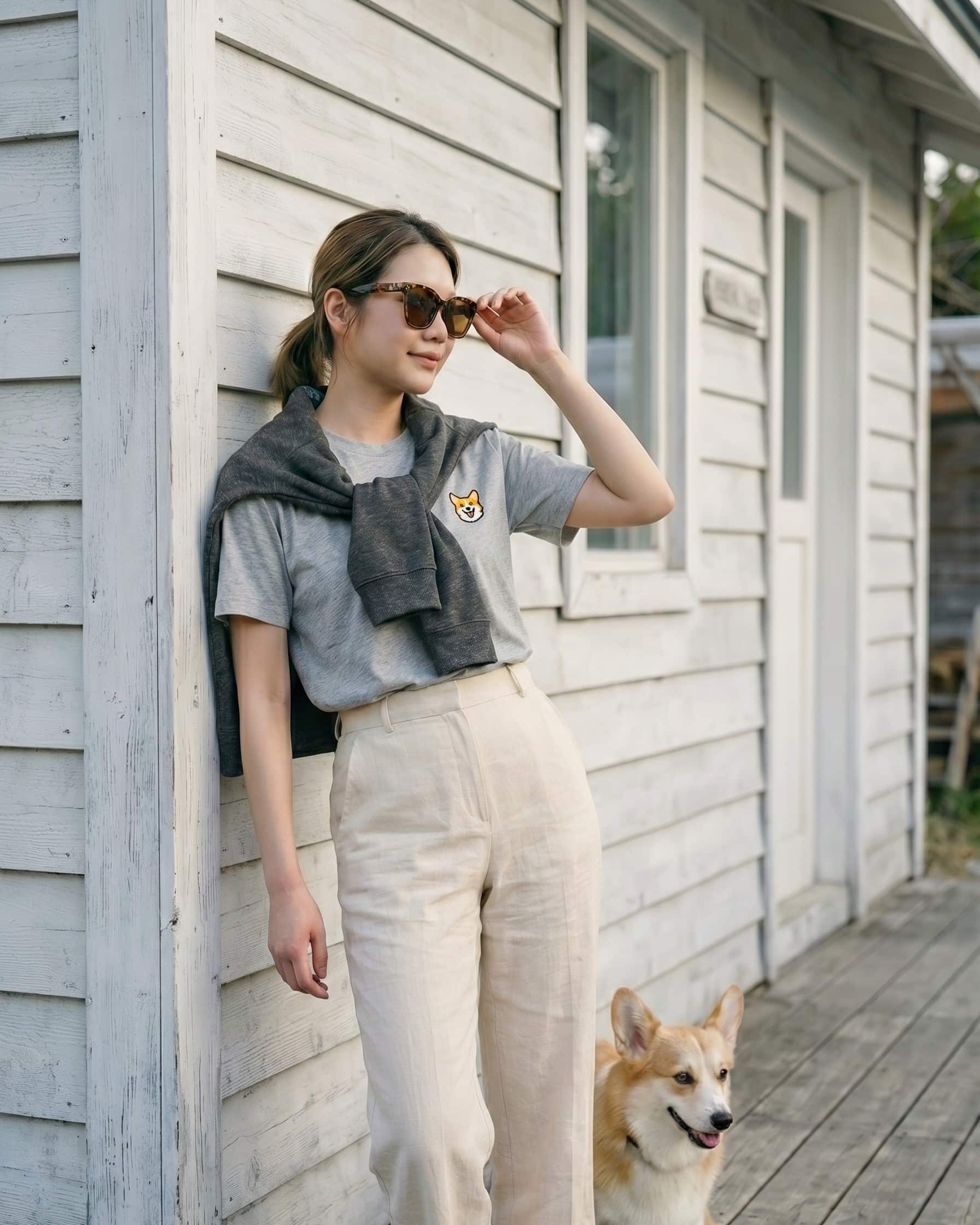 Woman standing outside wooden house wearing Light Grey Heather T-shirt with minimalist embroidered Corgi icon on left chest.