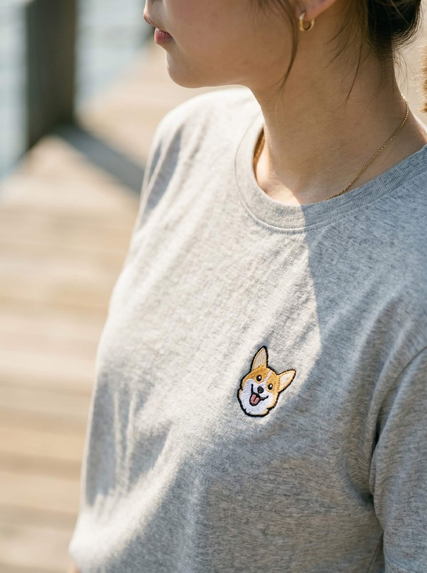 Close up of woman wearing Light Grey Heather T-shirt with minimalist embroidered Corgi icon on left chest.