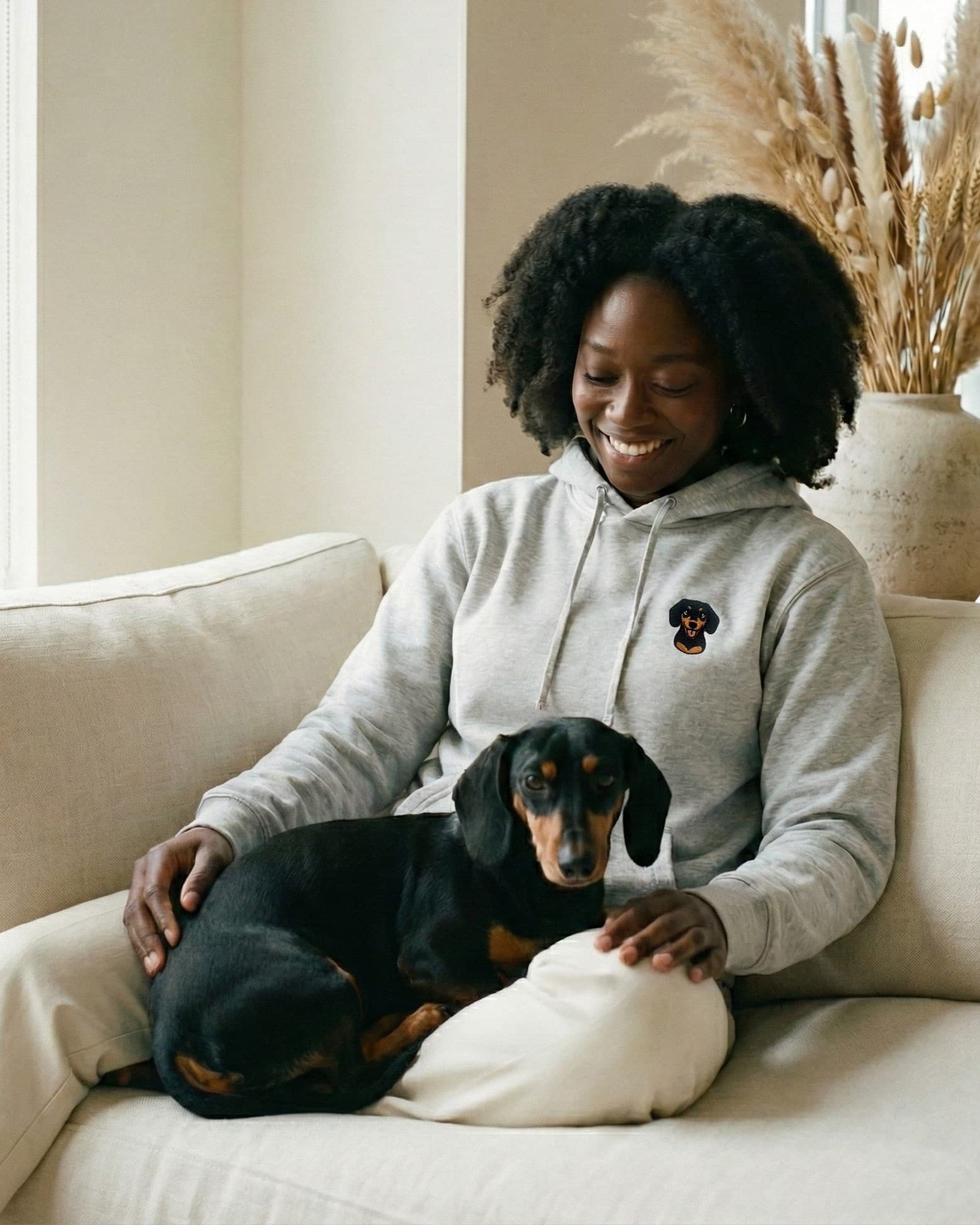 Woman sitting on couch with Dachshund in her lap, wearing Light Grey Heather Hoodie with minimalist embroidered Dachshund icon on left chest.