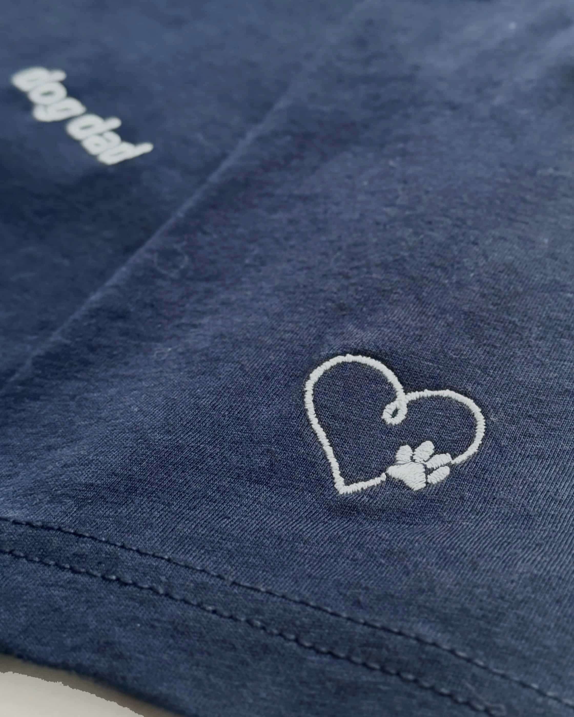 Close up of Navy t-shirt with minimalist embroidered phrase "dog dad" on left chest. Embroidered Heart and Paw icon on the left sleeve above cuff.