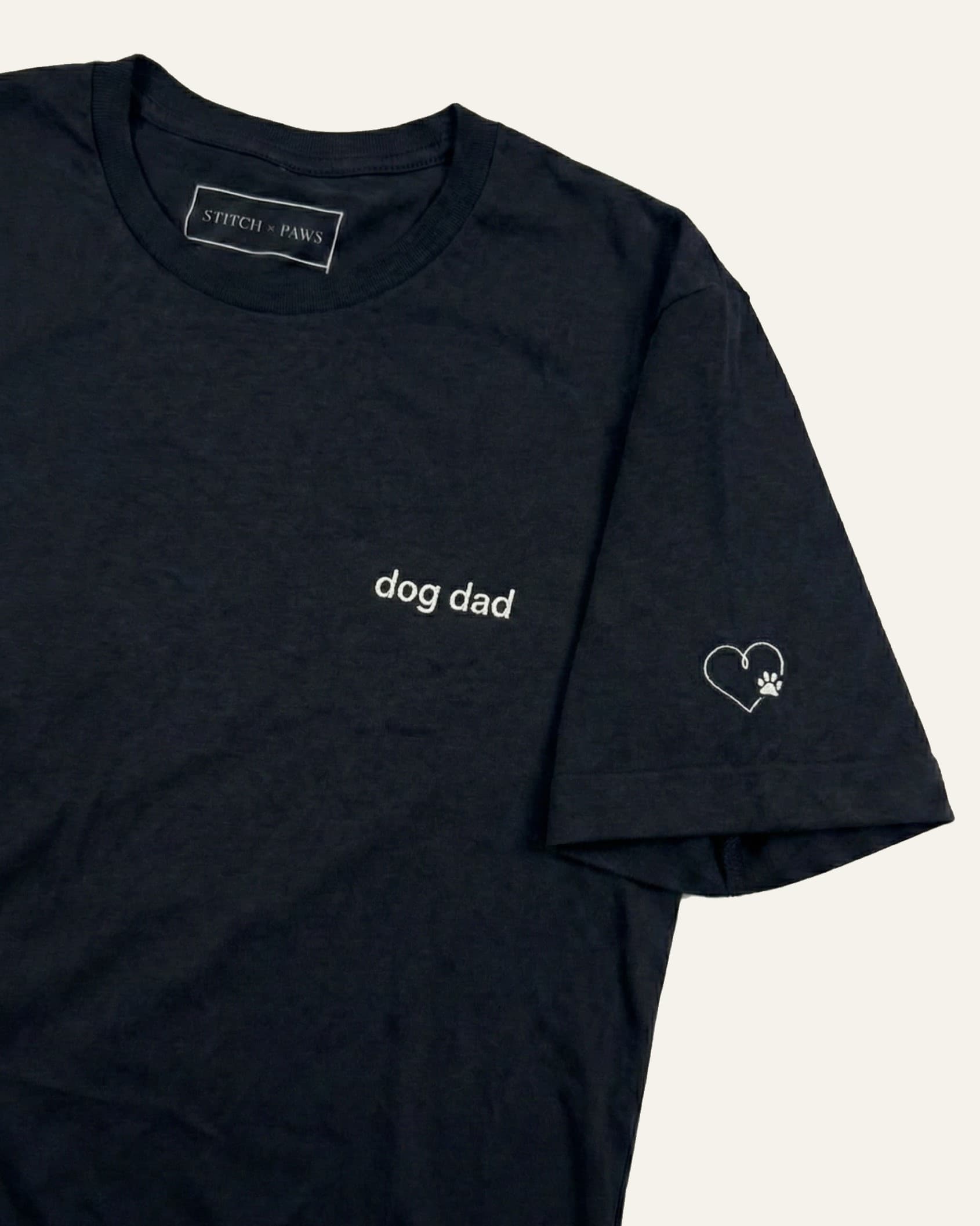 Black t-shirt with minimalist embroidered phrase "dog dad" on left chest. Embroidered Heart and Paw icon on the left sleeve above cuff.
