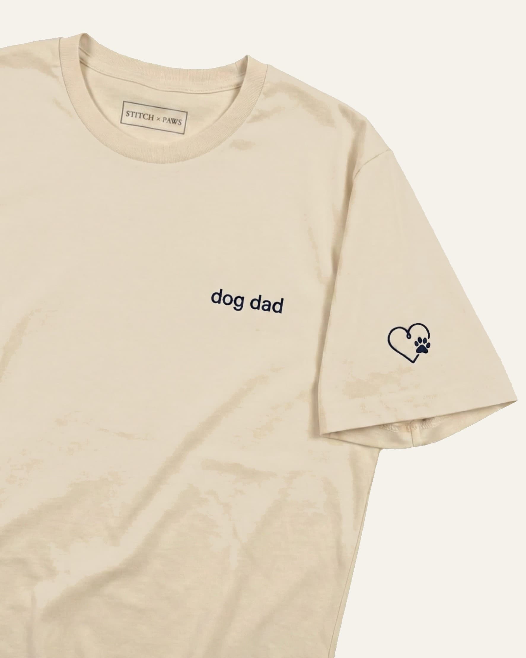 Beige t-shirt with minimalist embroidered phrase "dog dad" on left chest. Embroidered Heart and Paw icon on the left sleeve above cuff.