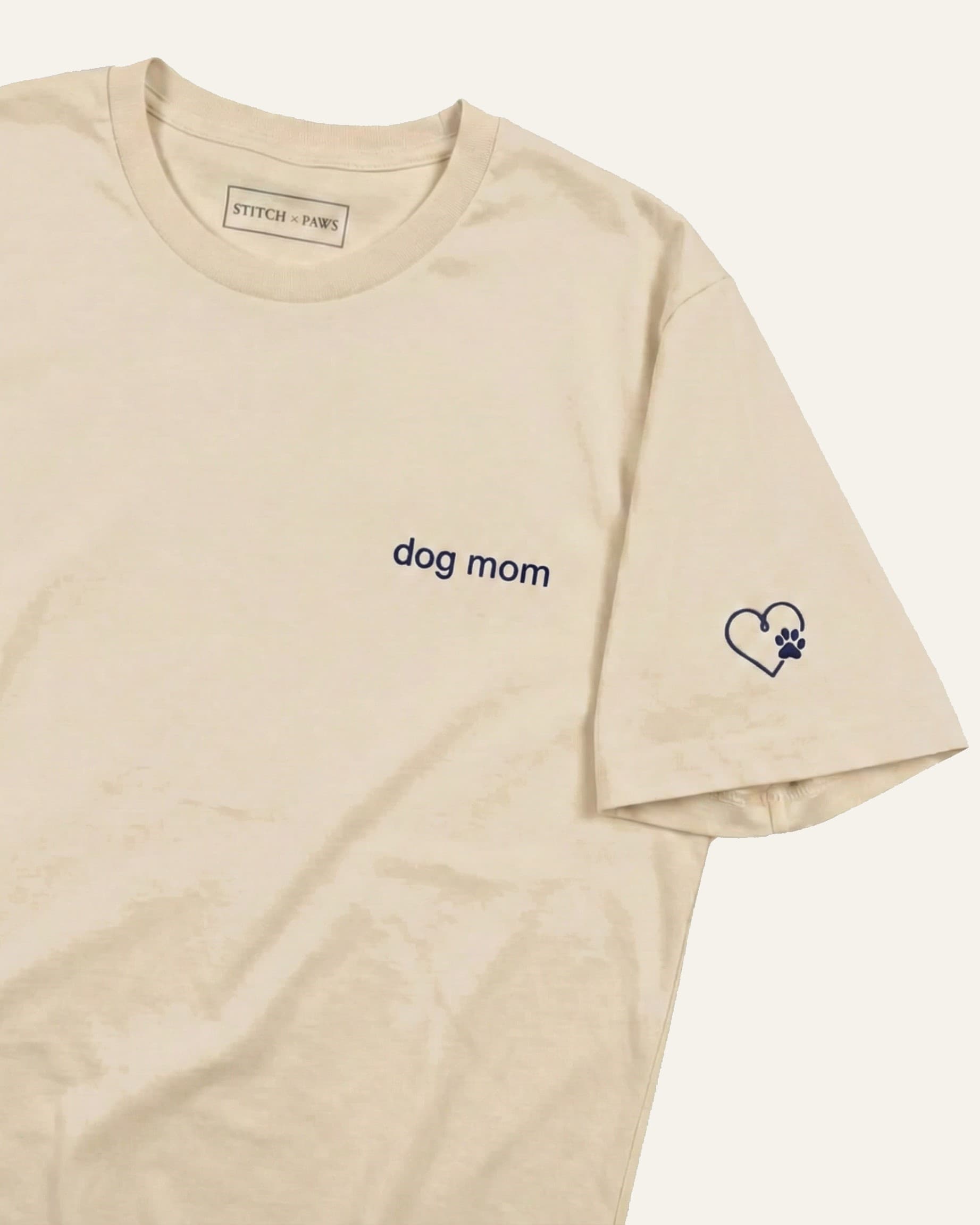 Beige t-shirt with minimalist embroidered phrase "dog mom" on left chest. Embroidered Heart and Paw icon on the left sleeve above cuff.