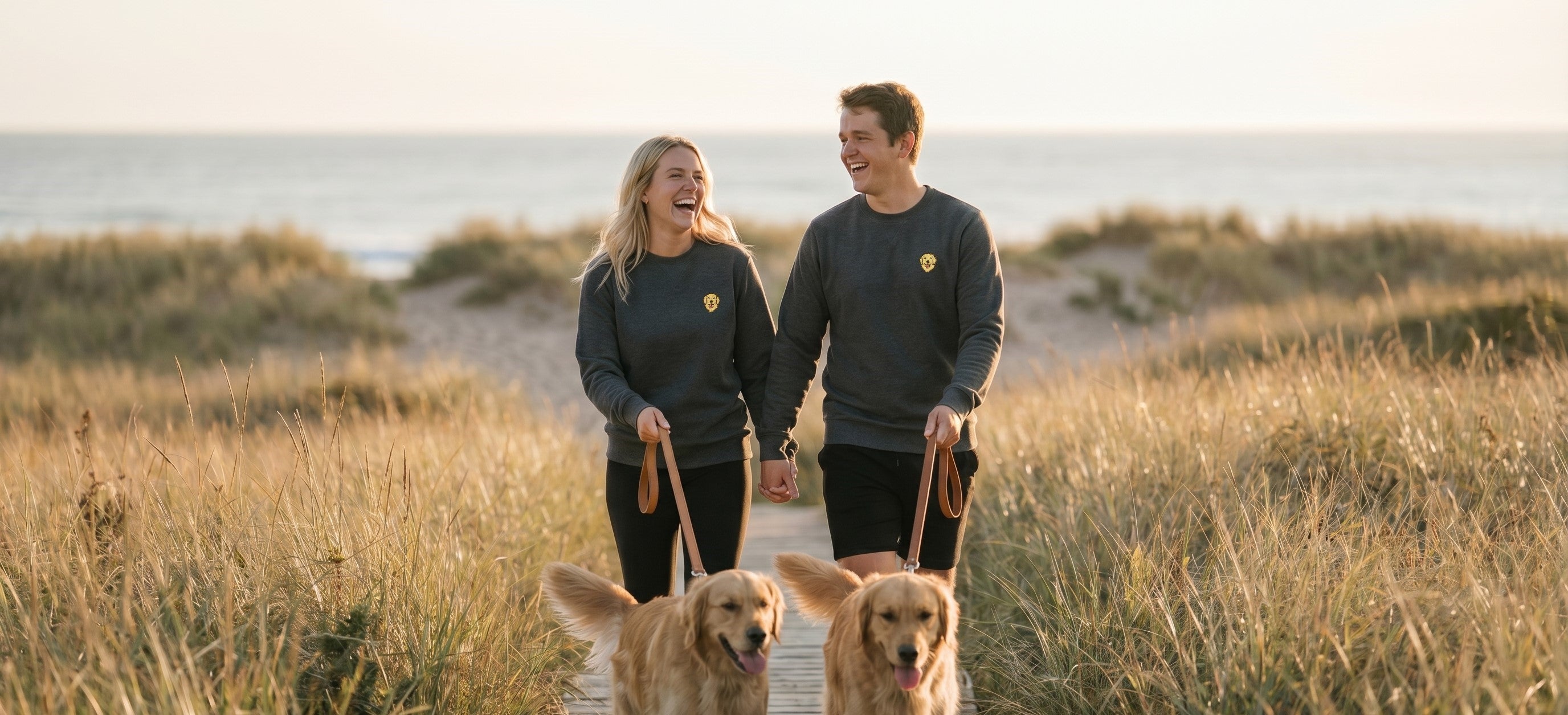 Man and woman walking two dogs on a wooden path by the beach.