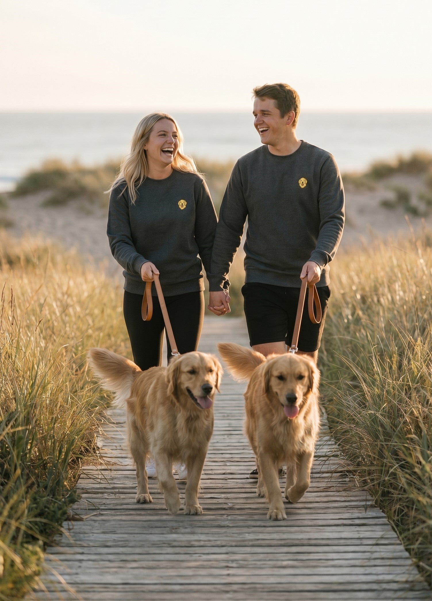 Man and woman walking two dogs on a wooden path by the beach.