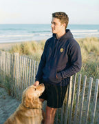 Man at the coast leaning against wooden fence with a Golden Retriever in front of him. He is wearing NavyHoodie with minimalist embroidered Golden Retriever icon on left chest.
