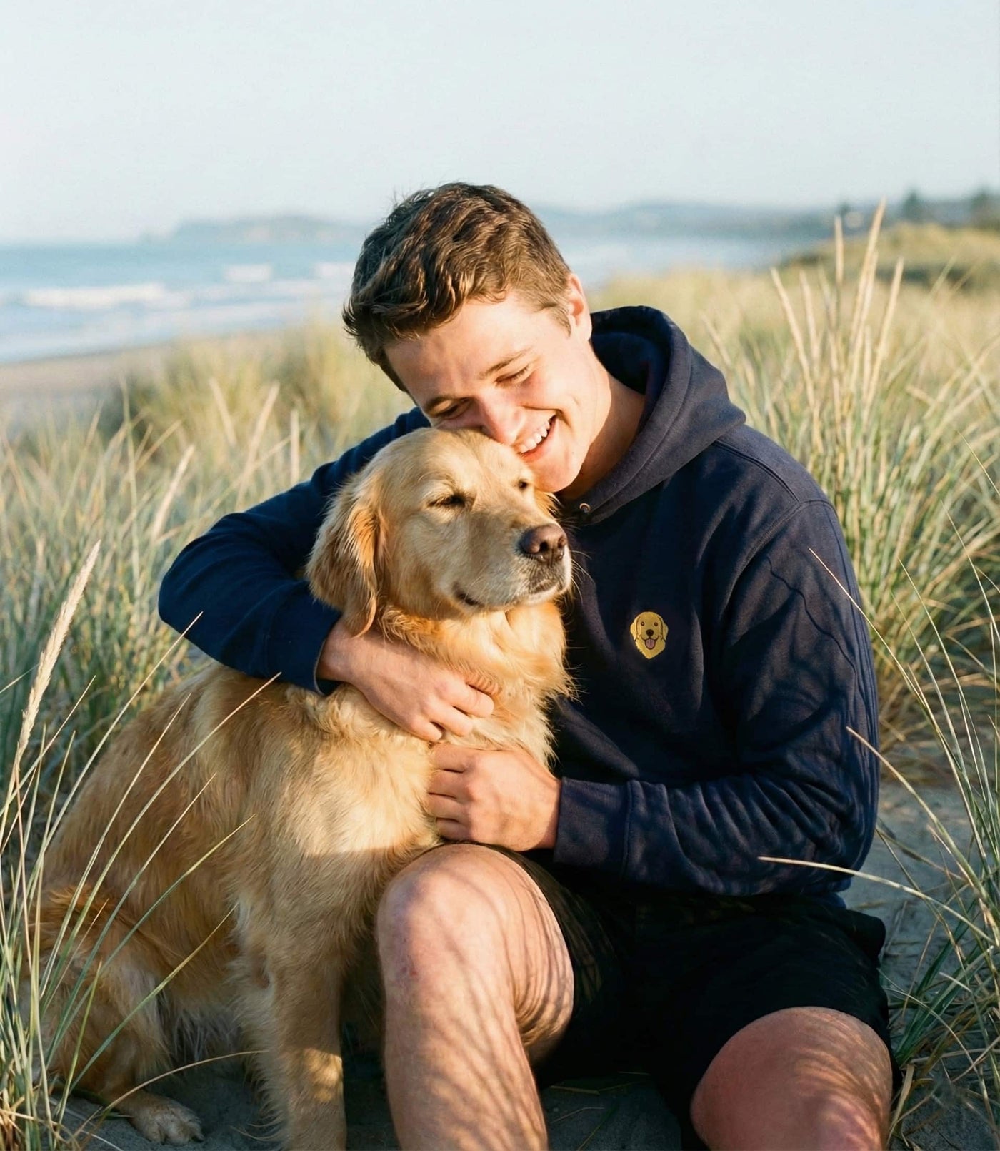 Man hugging golden retriever, wearing a Navy hoodie with minimalist embroidered Golden Retriever icon on left chest.