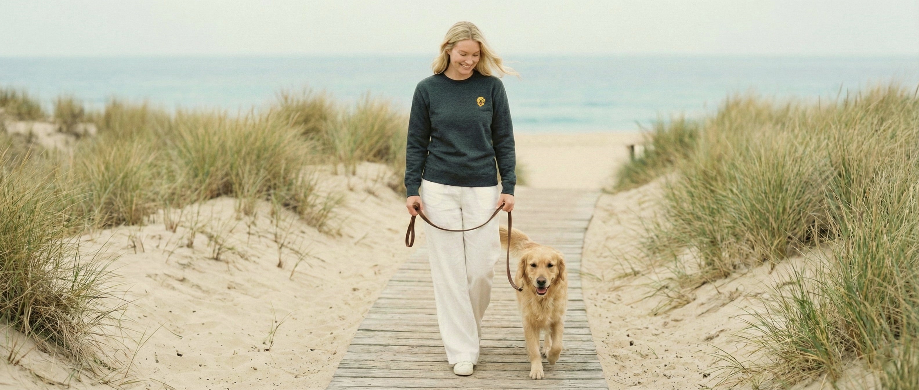 Woman walking with Golden Retriever on beach boardwalk, wearing Dark Grey Heather Sweatshirt with minimalist embroidered Golden Retriever icon on left chest.
