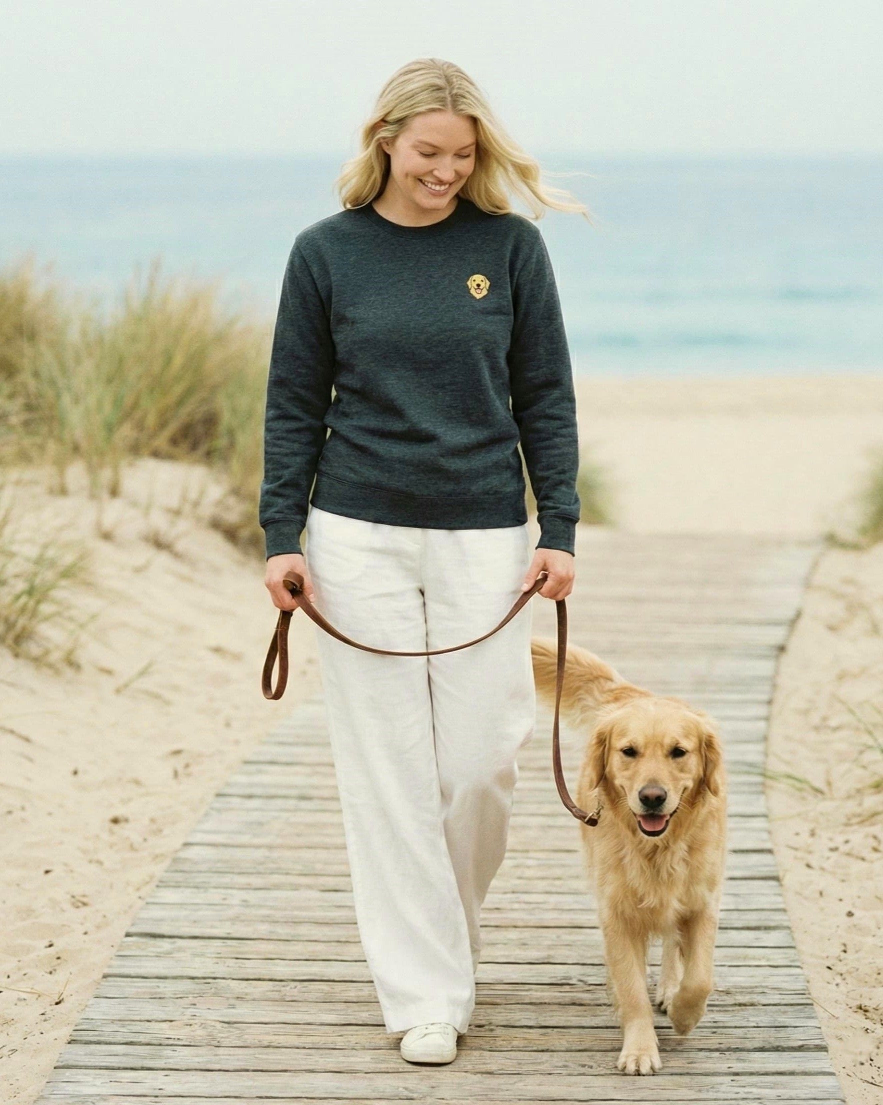 Woman walking with Golden Retriever on beach boardwalk, wearing Dark Grey Heather Sweatshirt with minimalist embroidered Golden Retriever icon on left chest.