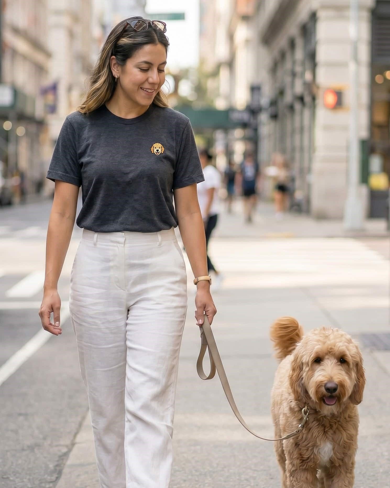 Woman walking Goldendoodle on city sidewalk, wearing Dark Grey Heather T-shirt with minimalist embroidered Goldendoodle icon on left chest.
