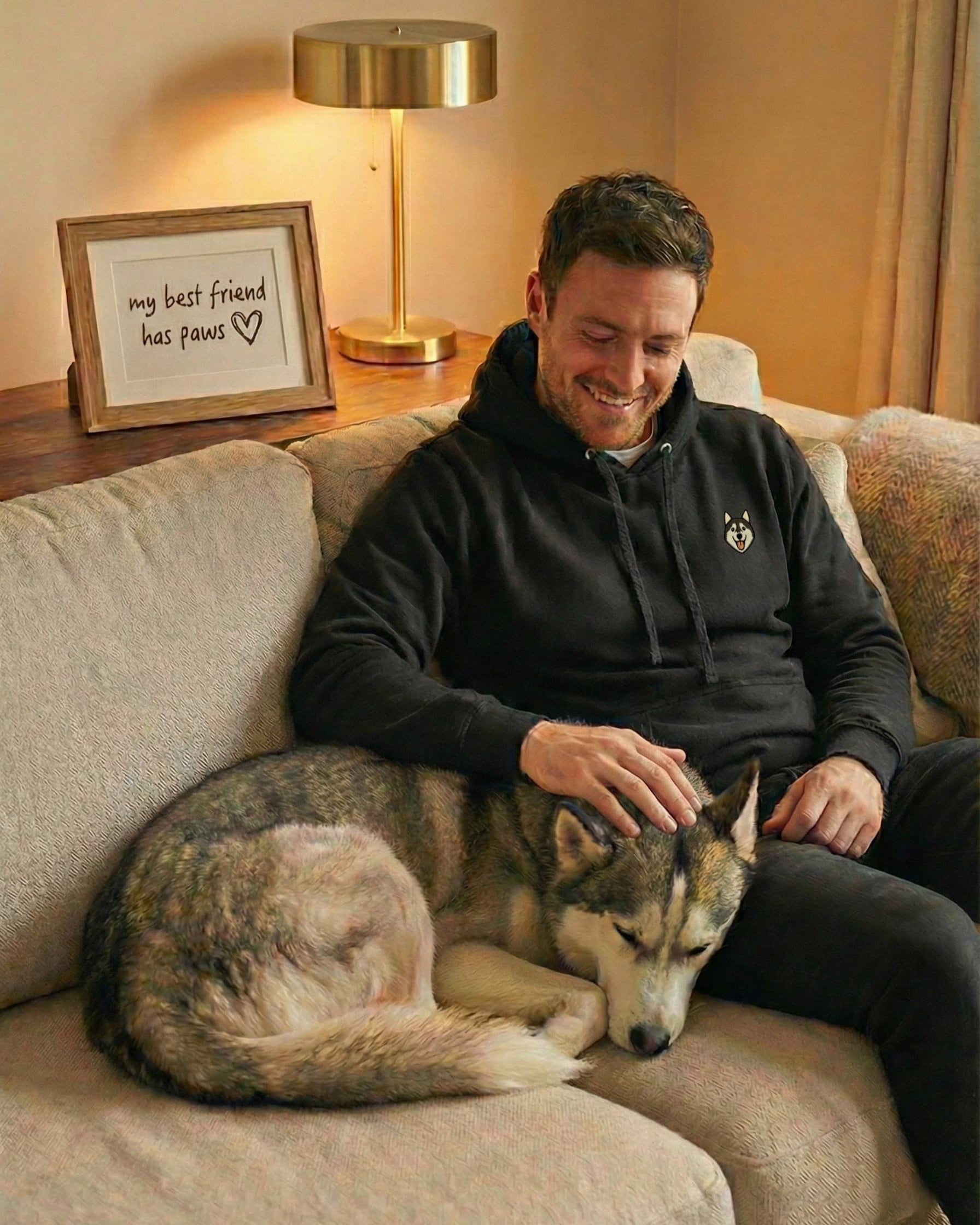 Man sitting on couch with Husky, wearing Black Hoodie with minimalist embroidered Husky icon on left chest.