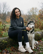 Woman sitting beside Husky, wearing Dark Grey Heather Sweatshirt with minimalist embroidered Husky icon on left chest.