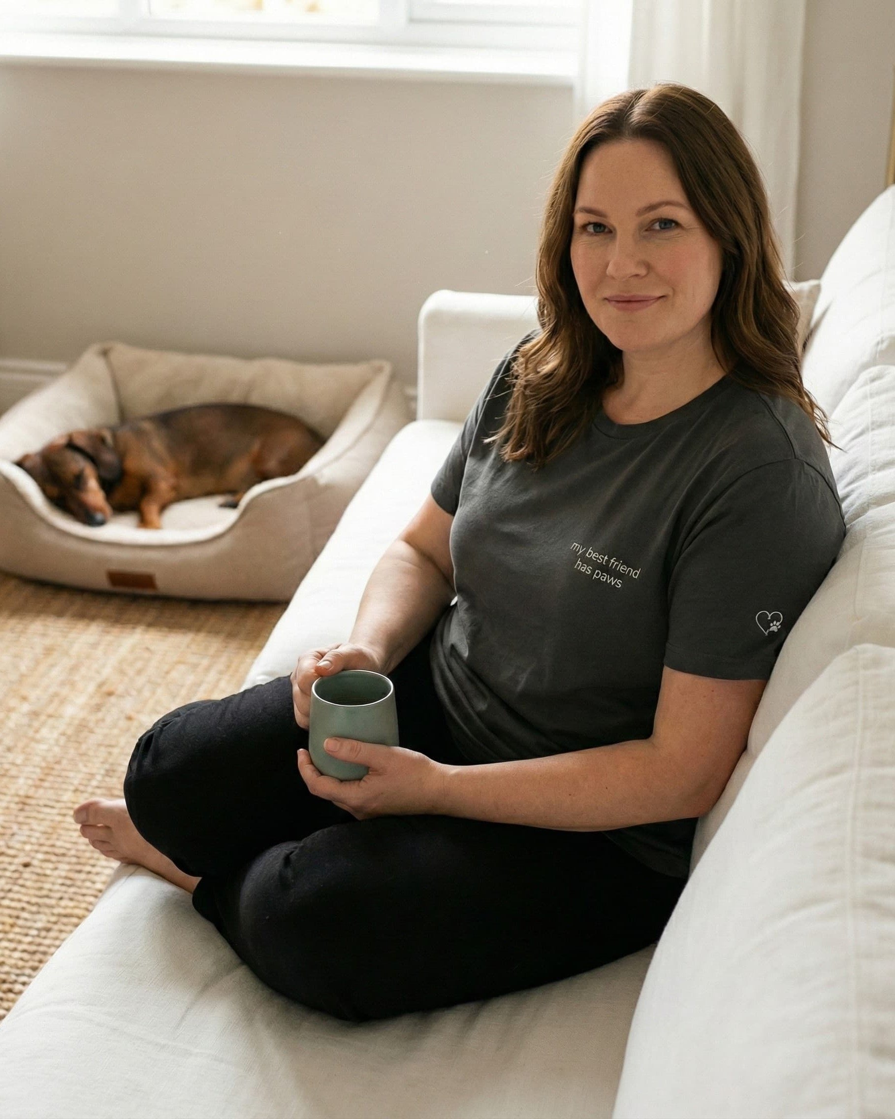 Woman sitting on couch, wearing Asphalt t-shirt with minimalist embroidered phrase "my best friend has paws" on left chest. Embroidered Heart and Paw icon on the left sleeve above cuff.