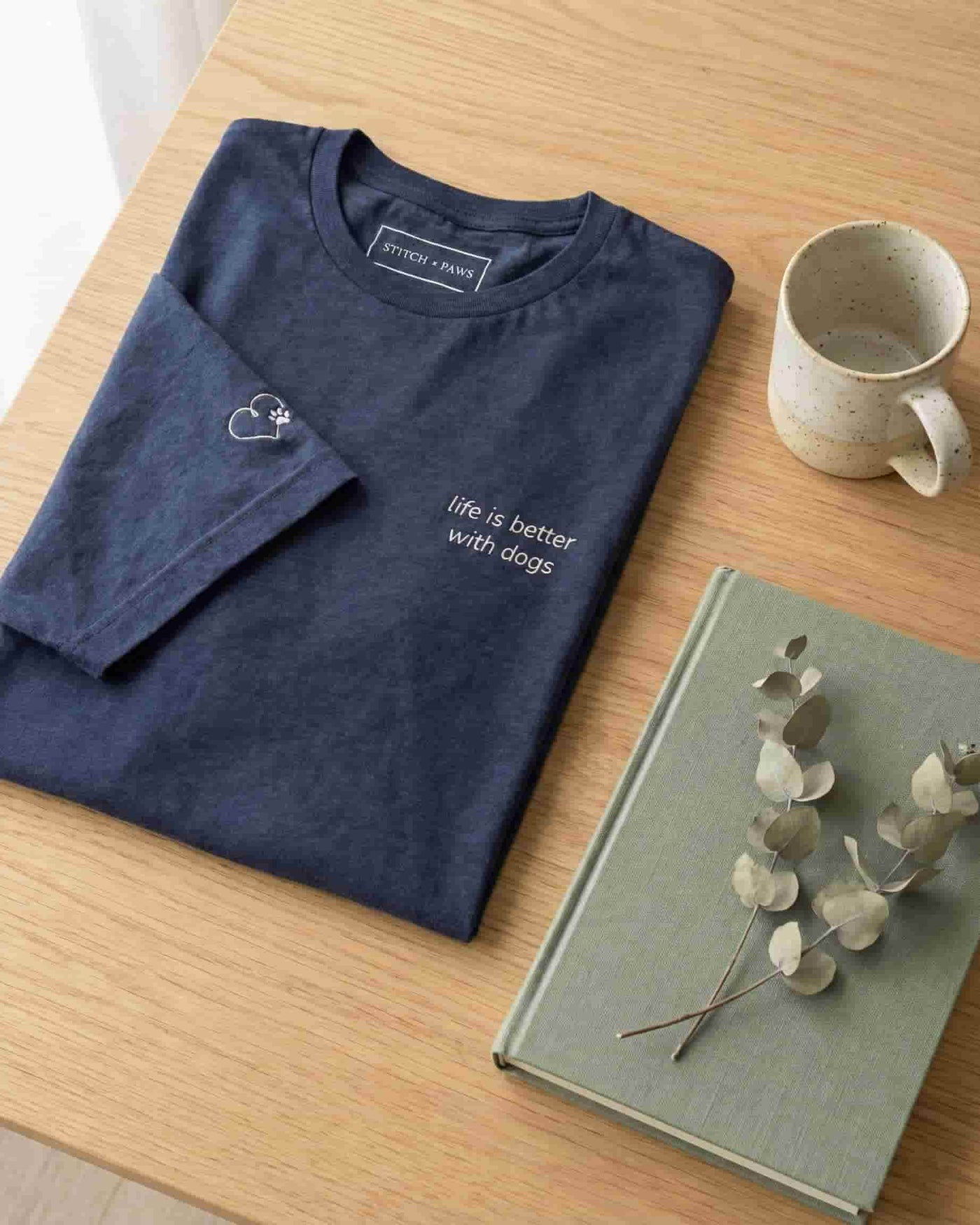 Folded Navy t-shirt with minimalist embroidered phrase "life is better with dogs" on left chest. Embroidered Heart and Paw icon on the left sleeve above cuff.